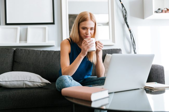 Thoughtful pretty young woman drinking coffee and using laptop at home